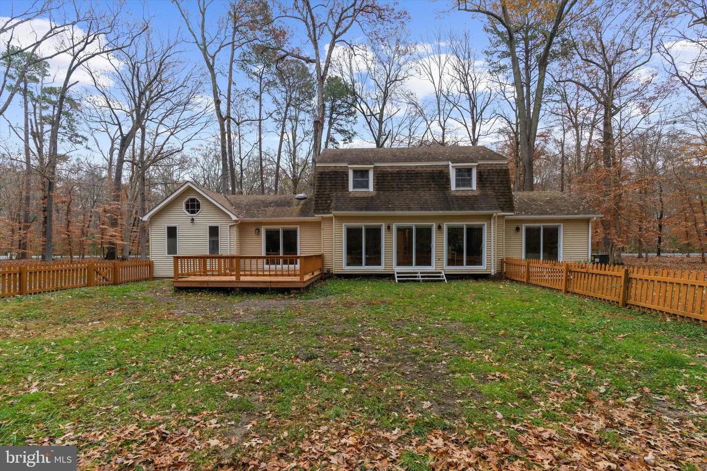 Backyard view of a tan house with a brown roof, a wooden deck, and a wooden fence, surrounded by trees and fallen leaves.