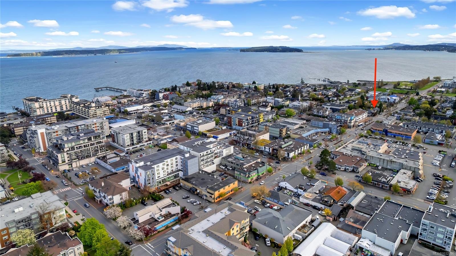 Aerial view of a coastal town with buildings, streets, cars, and a body of water in the background under a blue sky with clouds. An arrow points to a specific building.