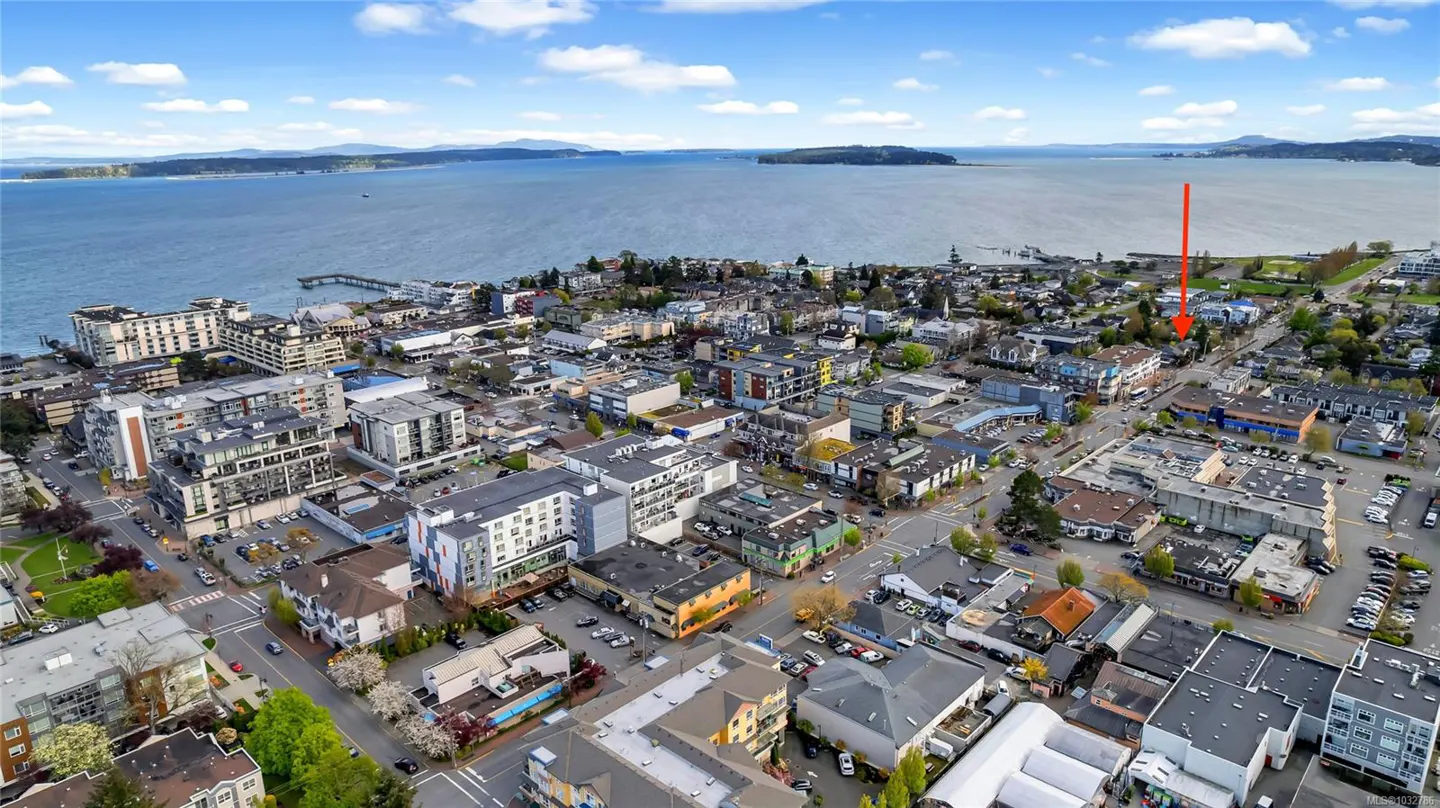 Aerial view of a coastal town with buildings, streets, cars, and a body of water in the background under a blue sky with clouds. An arrow points to a specific building.