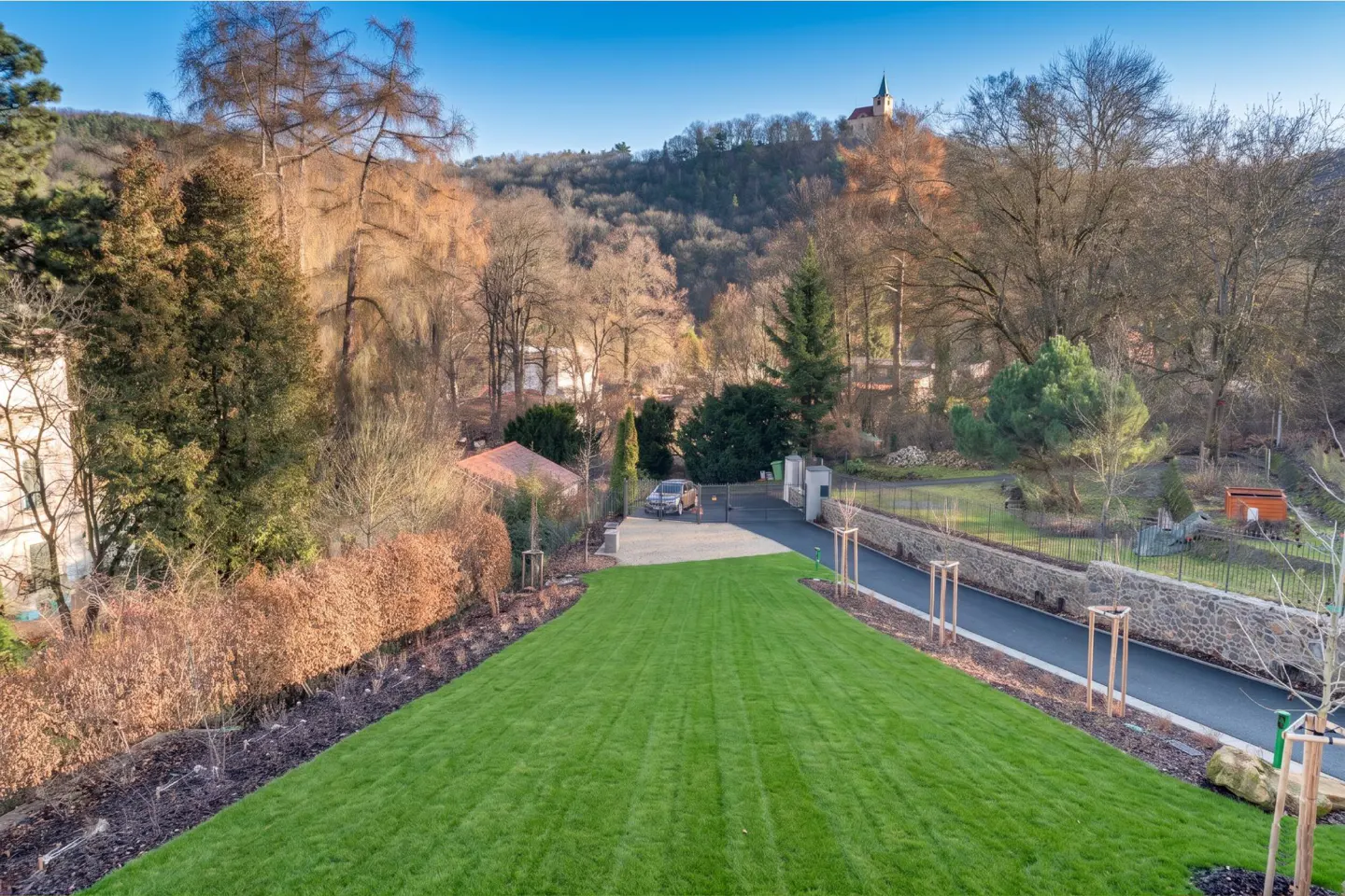 View of a green lawn leading to a driveway with a car, trees, and a building on a hill.