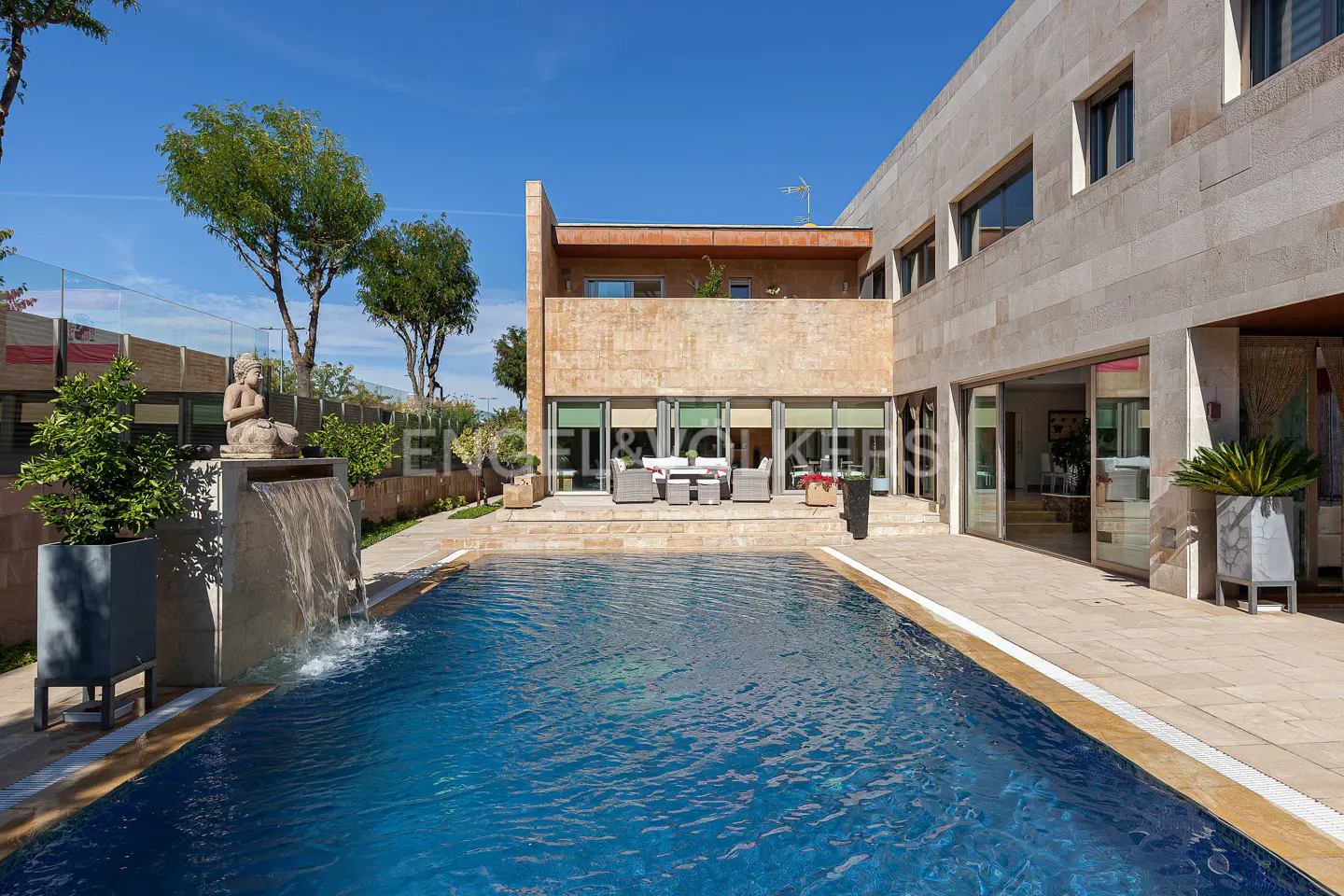Modern stone villa with a blue swimming pool, a water feature with a Buddha statue, and a patio under a clear blue sky.