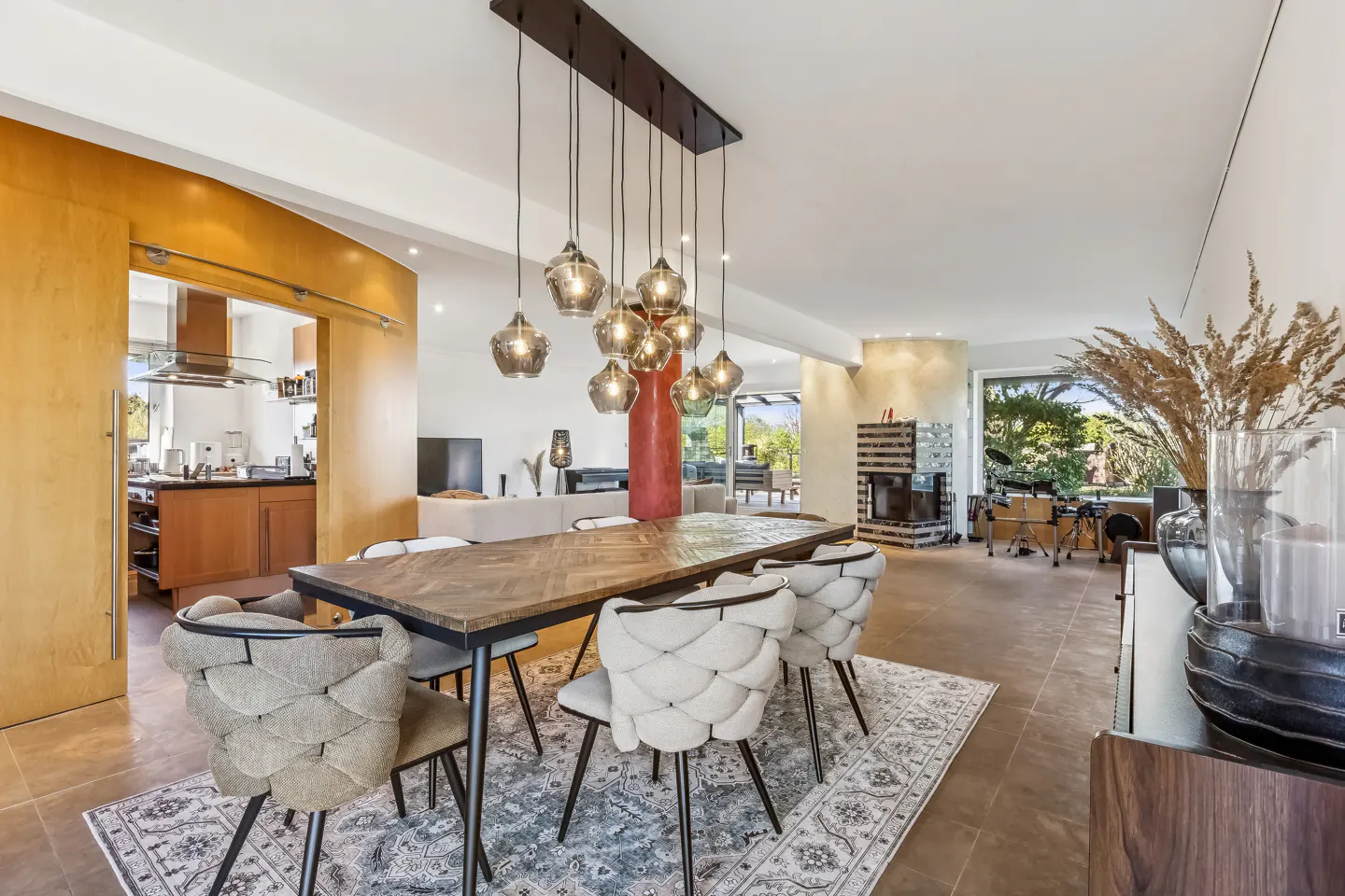 Open-concept dining room with a long wooden table, upholstered chairs, and a modern chandelier. Kitchen and living room are visible in the background.