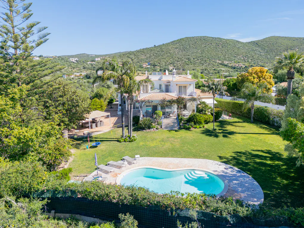 Aerial view of a white villa with a pool, green lawn, palm trees, and mountain backdrop under a clear blue sky.