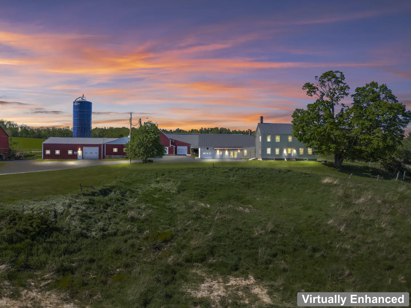 A farm house with red barns and a blue silo sits on a green lawn under a colorful sunset sky.