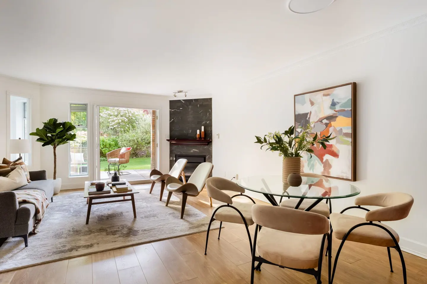 Bright living room with a gray sofa, glass table with four chairs, and a marble fireplace. Open doors lead to a green garden.