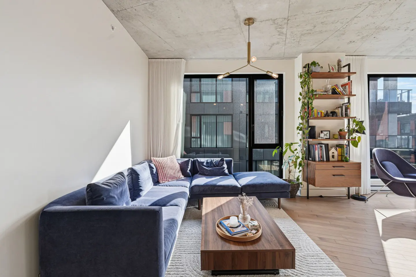 Bright living room with a blue sectional sofa, wood coffee table, and a bookshelf with plants near a window.