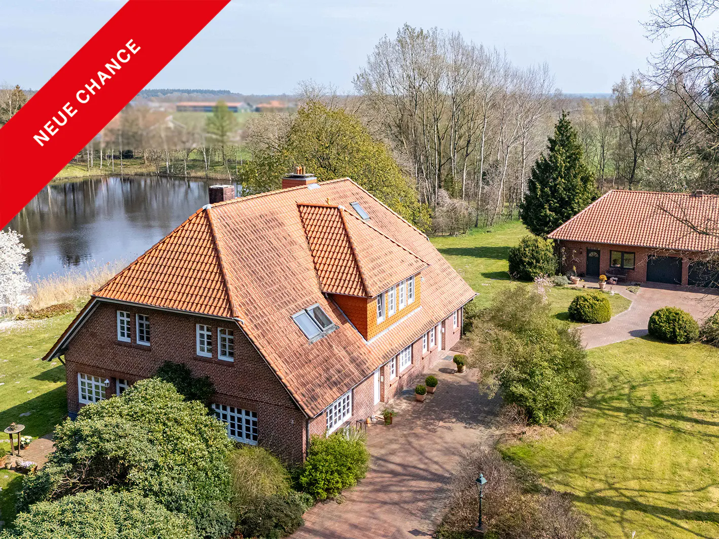 Aerial view of a brick house with a red tile roof, a lake, and a red "Neue Chance" banner.