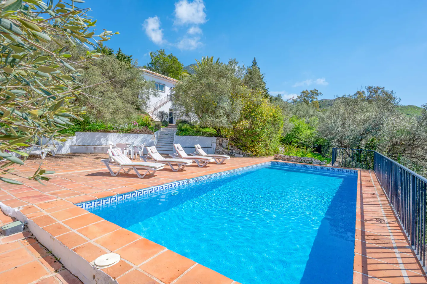 Outdoor pool with blue water, surrounded by terracotta tiles and lounge chairs. A white house is visible in the background.