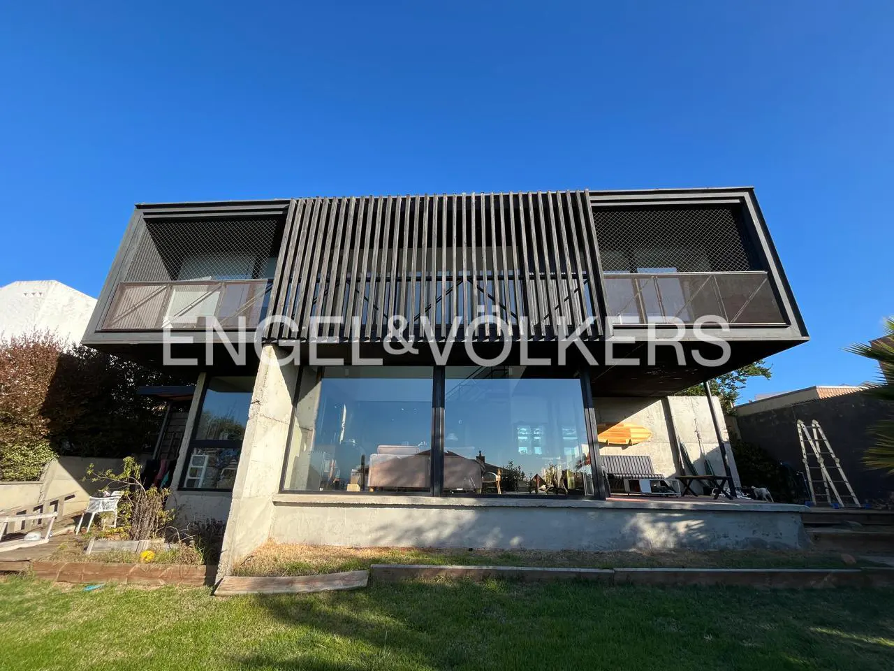 Exterior view of a modern two-story house with large windows and a gray facade, set against a clear blue sky. The Engel & Volkers logo is visible.