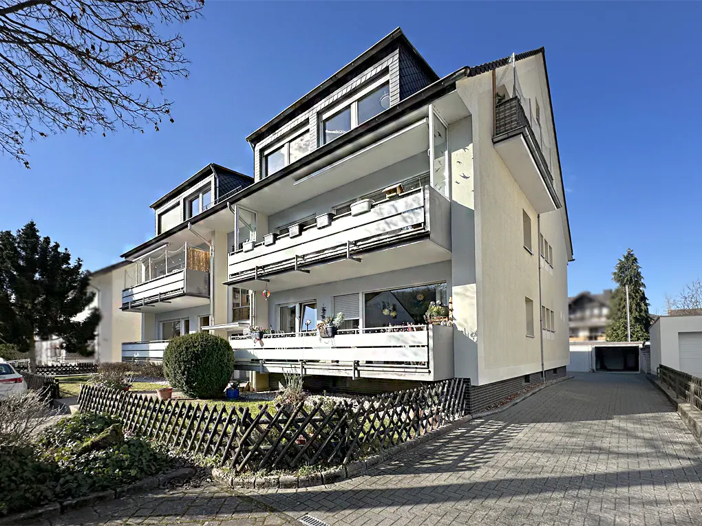 Exterior view of a three-story apartment building with white balconies and a gray roof on a sunny day.