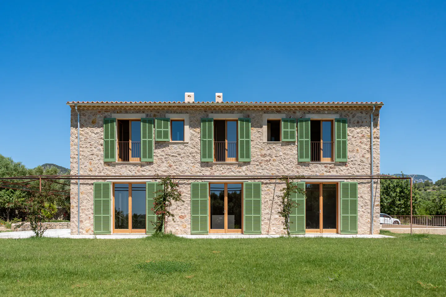 Two-story stone house with green shutters and a green lawn under a clear blue sky.