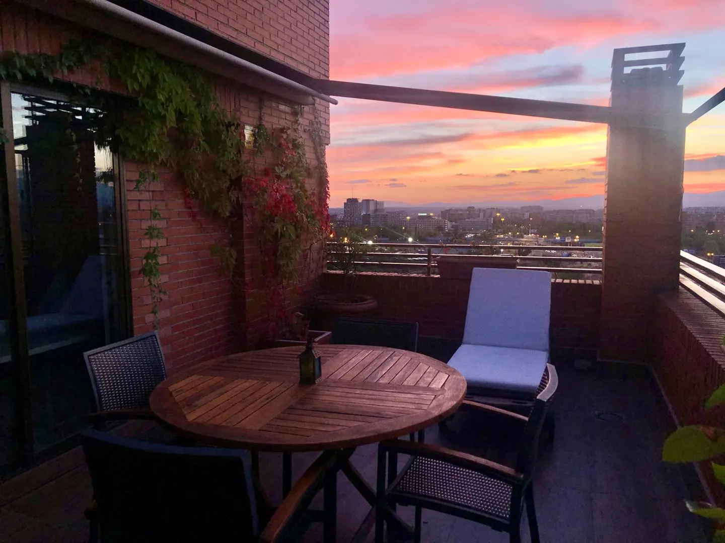 Rooftop patio with a round wooden table, chairs, and a white lounge chair overlooking a city skyline at sunset. Brick wall with vines.