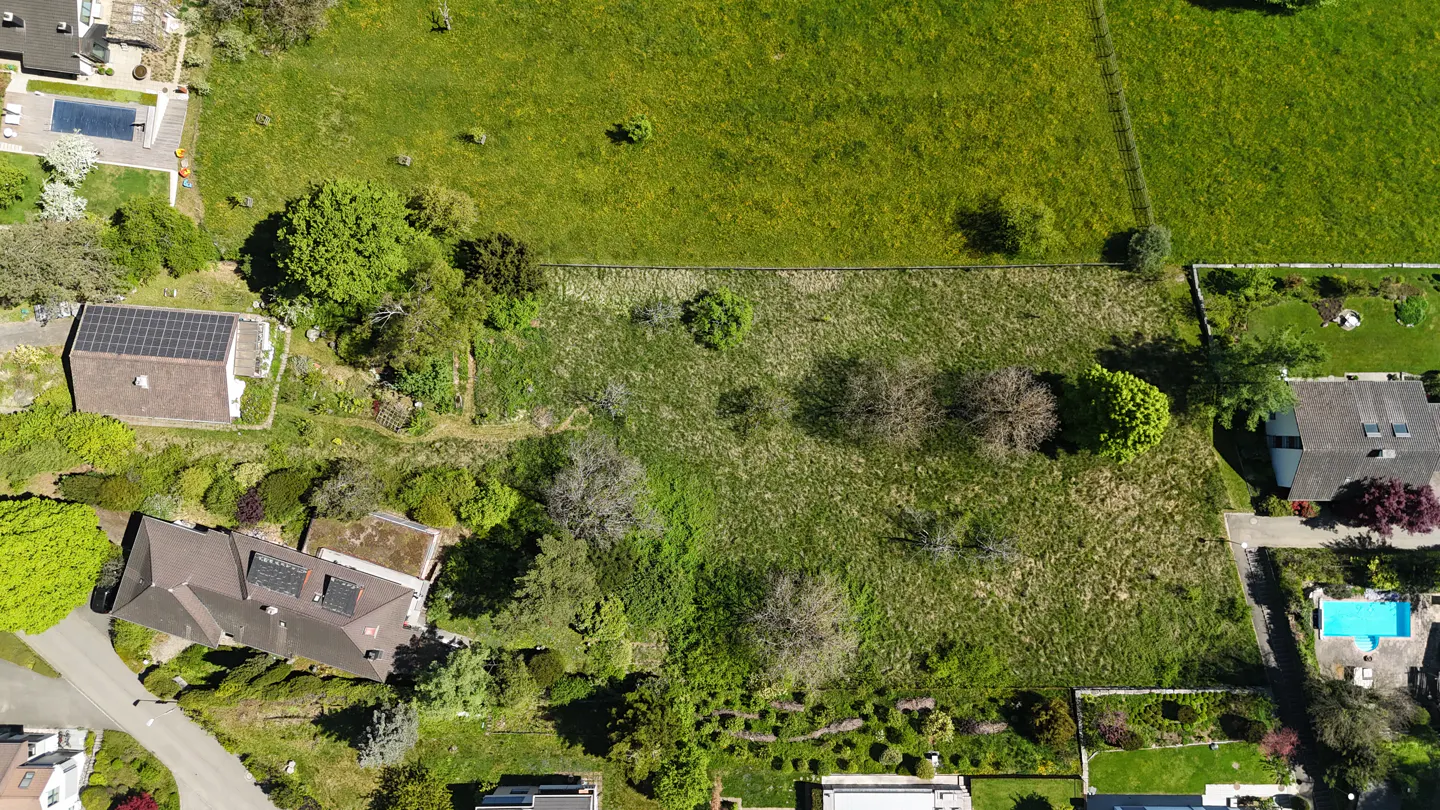 Aerial view of a grassy lot surrounded by houses, trees, and a green field.