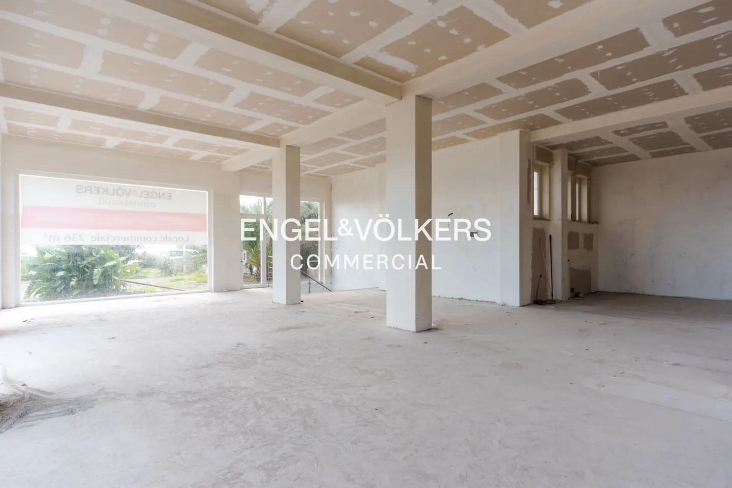 An unfinished commercial space with white walls, concrete floors, and exposed ceiling panels. Large windows provide natural light.