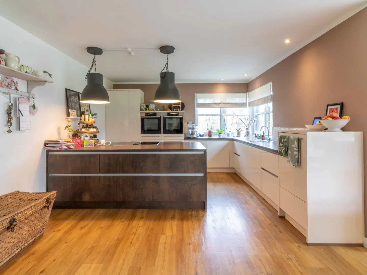 Bright kitchen with wood floors, a brown island with black pendant lights, and white cabinets. A wicker chest sits in the foreground.