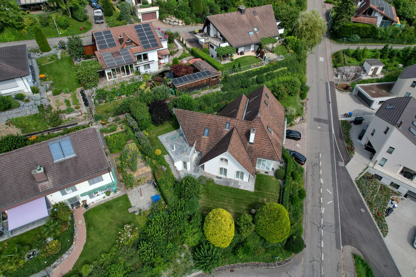 Aerial view of a white house with a brown roof, surrounded by lush greenery and other houses in a residential neighborhood.