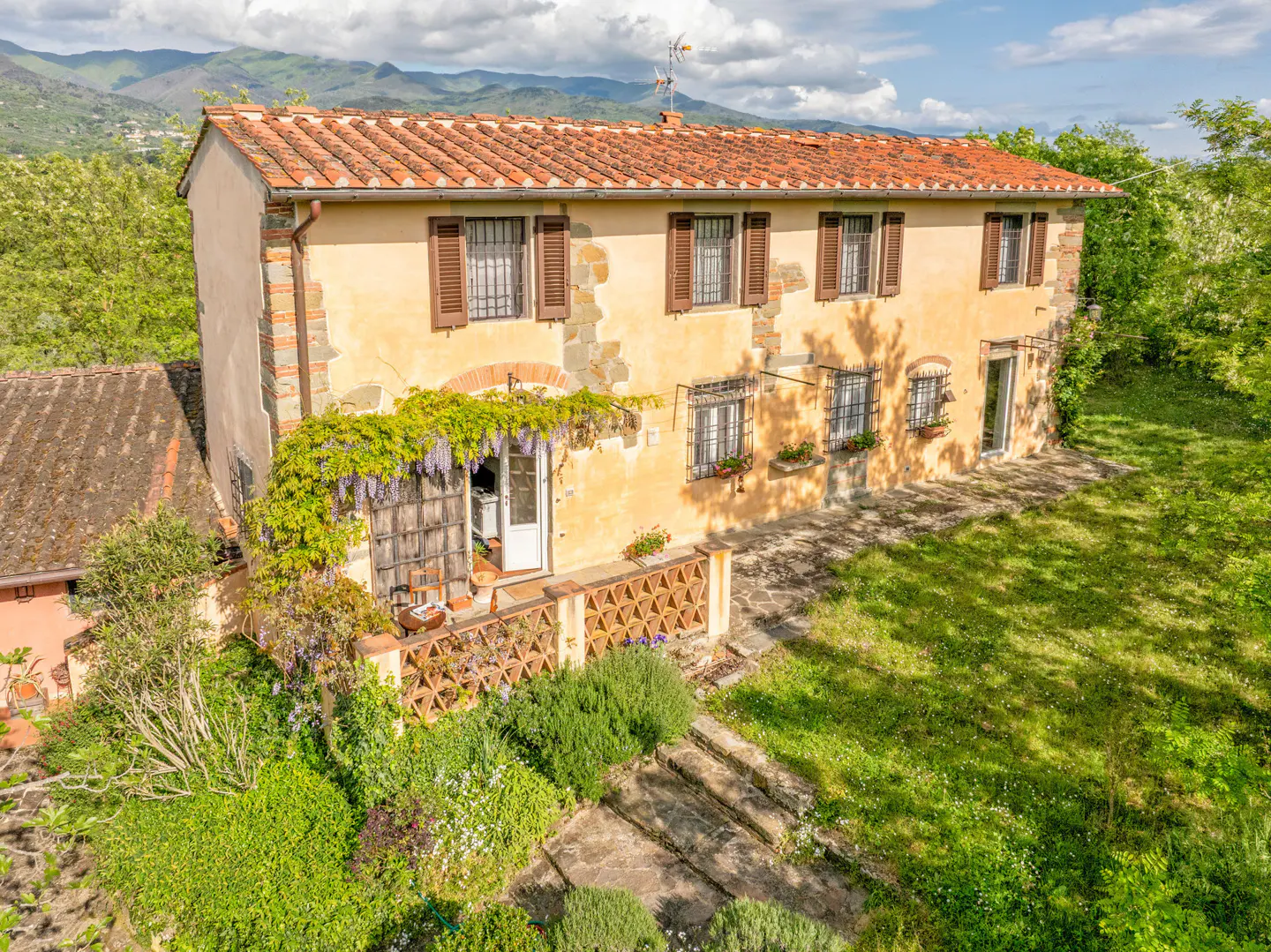 Two-story yellow house with a red tile roof, brown shutters, and a stone patio with green plants. Mountains are in the background.