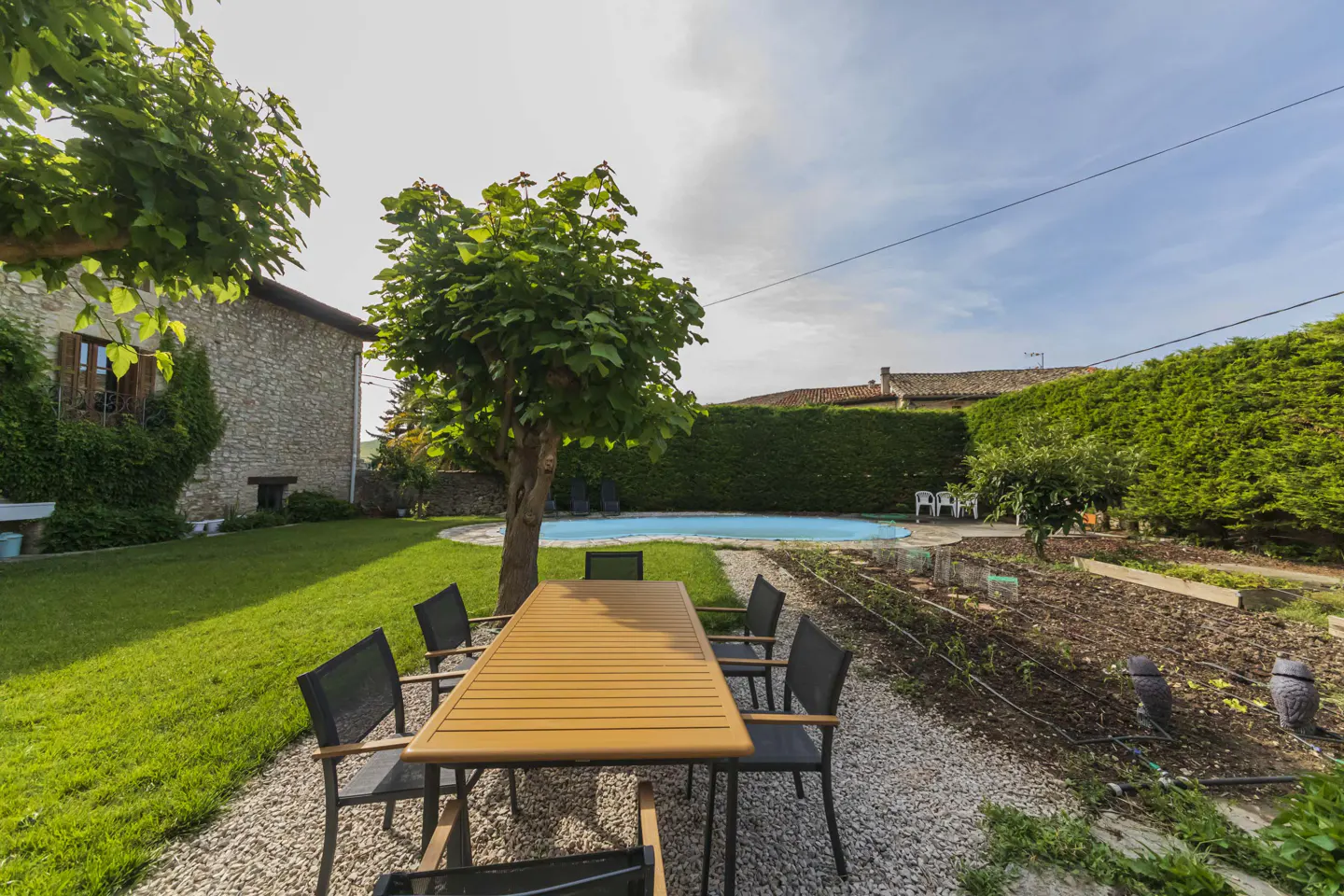 Outdoor dining area with a wooden table and chairs on a gravel patio, overlooking a green lawn, pool, and garden.