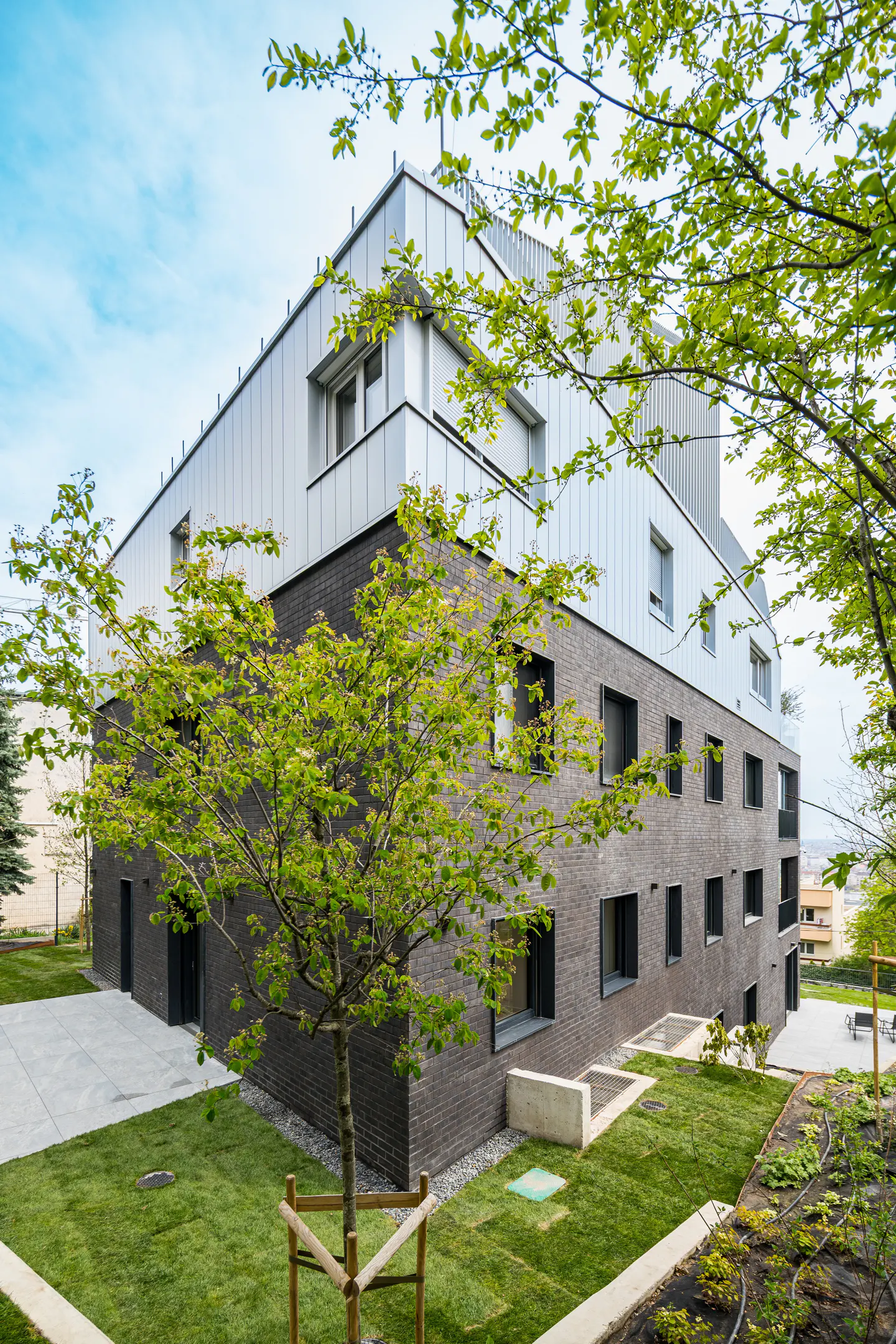 Modern building with a gray brick base and silver metal top, surrounded by green grass and trees under a blue sky.
