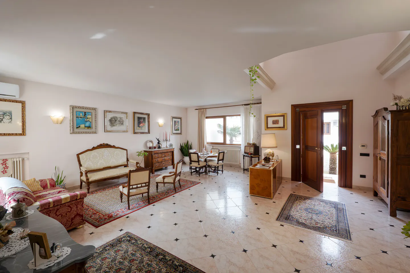 A bright, spacious living room with antique furniture, framed art, and a dining table near a window. The floor is tiled with black stars.