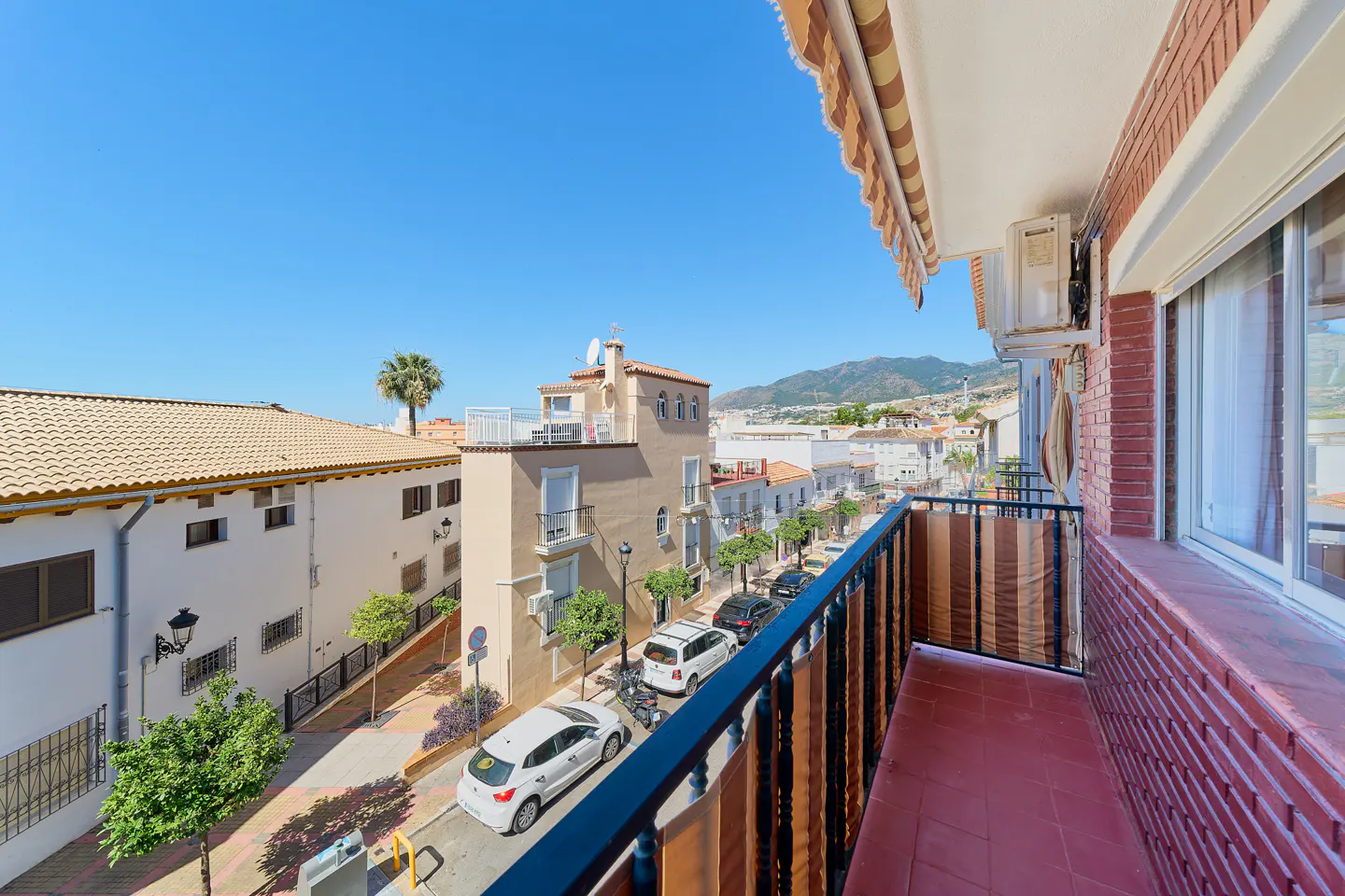 View from a balcony with red tiled floors overlooking a sunny street with parked cars, white buildings, and distant hills.