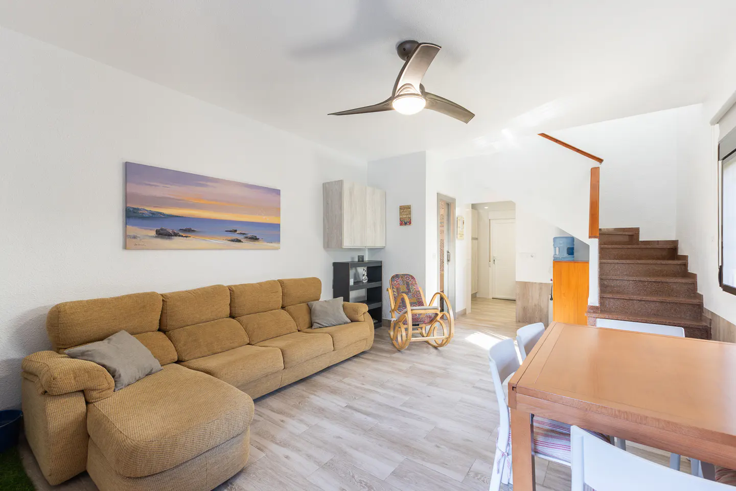 Living room with a brown sectional sofa, a painting of a beach, and a wooden dining table with white chairs.
