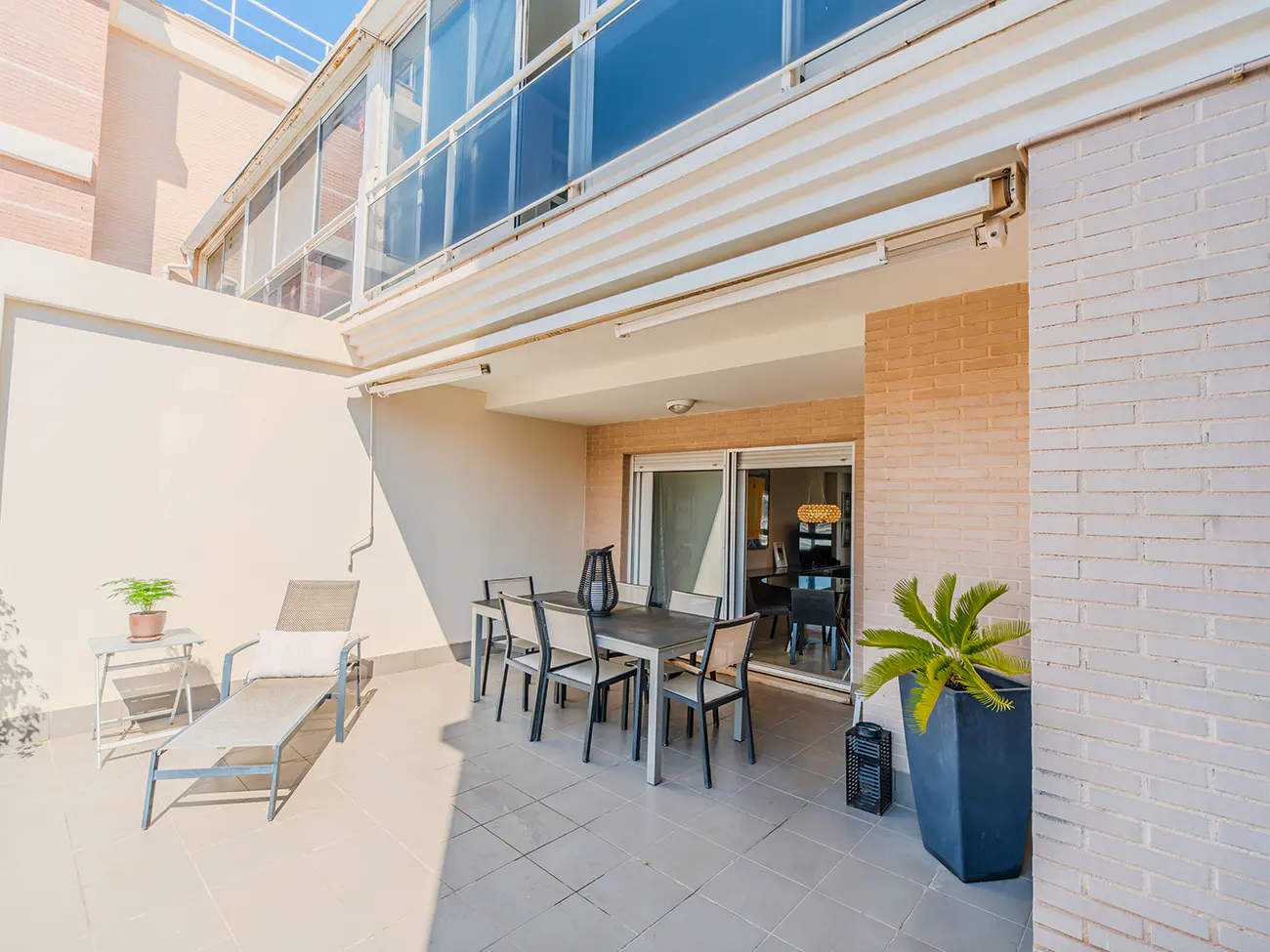 A sunny outdoor patio with a dining set, a lounge chair, a potted plant, and sliding glass doors leading to the interior.