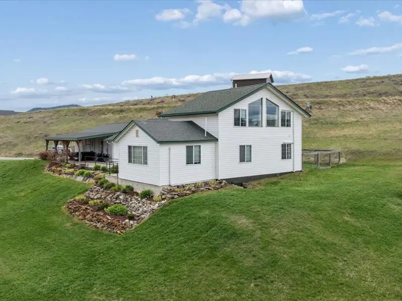 White two-story house with a green roof on a grassy hill under a blue sky with clouds.