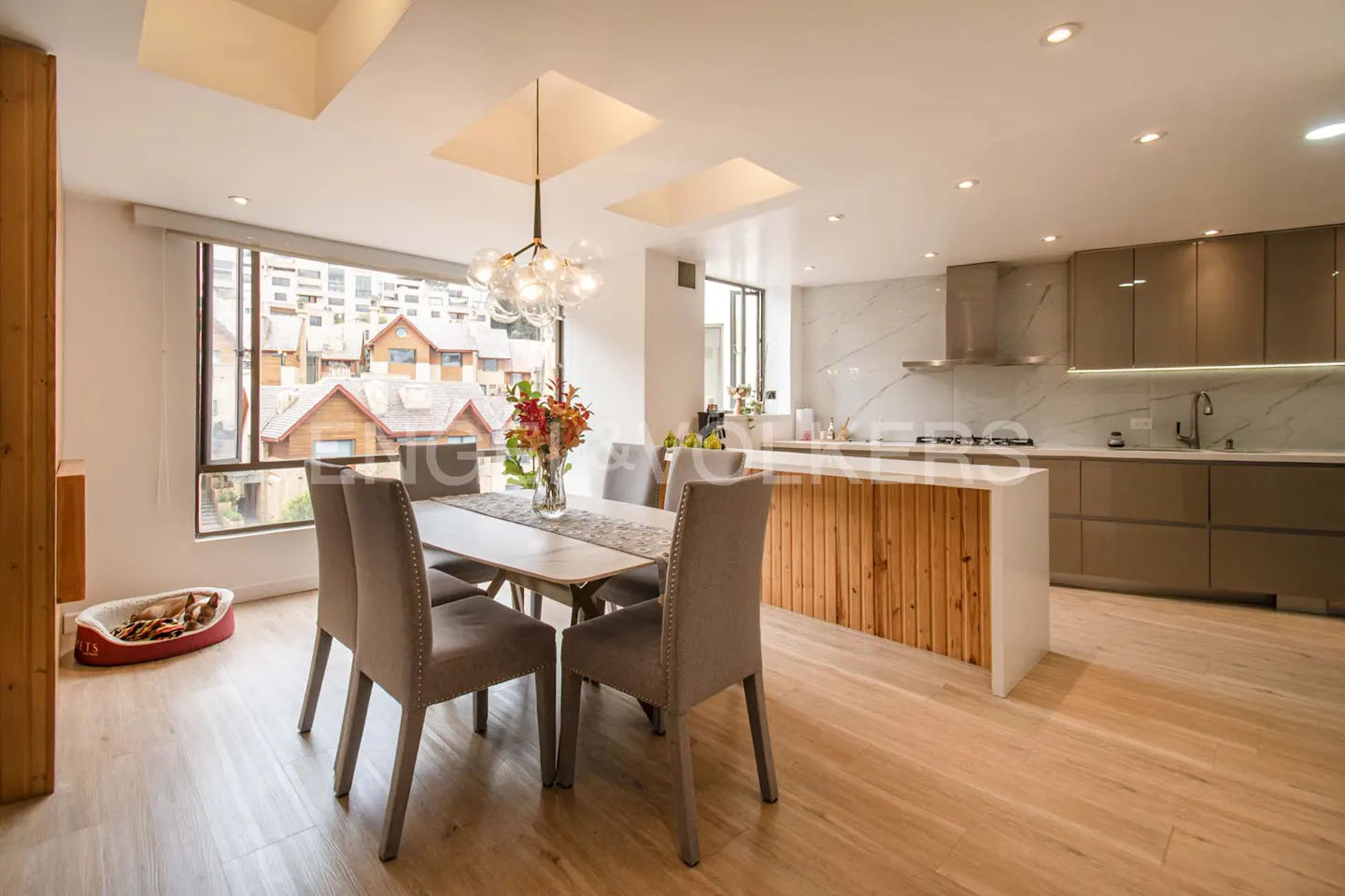 Bright, modern kitchen and dining area with wood floors, gray chairs, and a view of houses outside the window. A dog bed sits near the window.