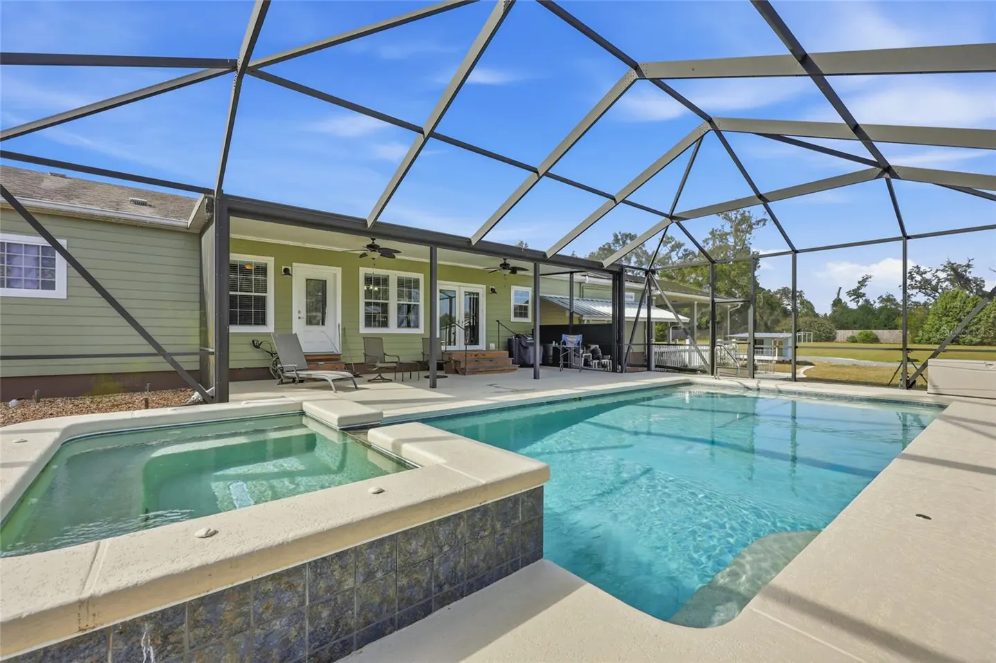 A screened-in pool and hot tub with a green house in the background under a blue sky.