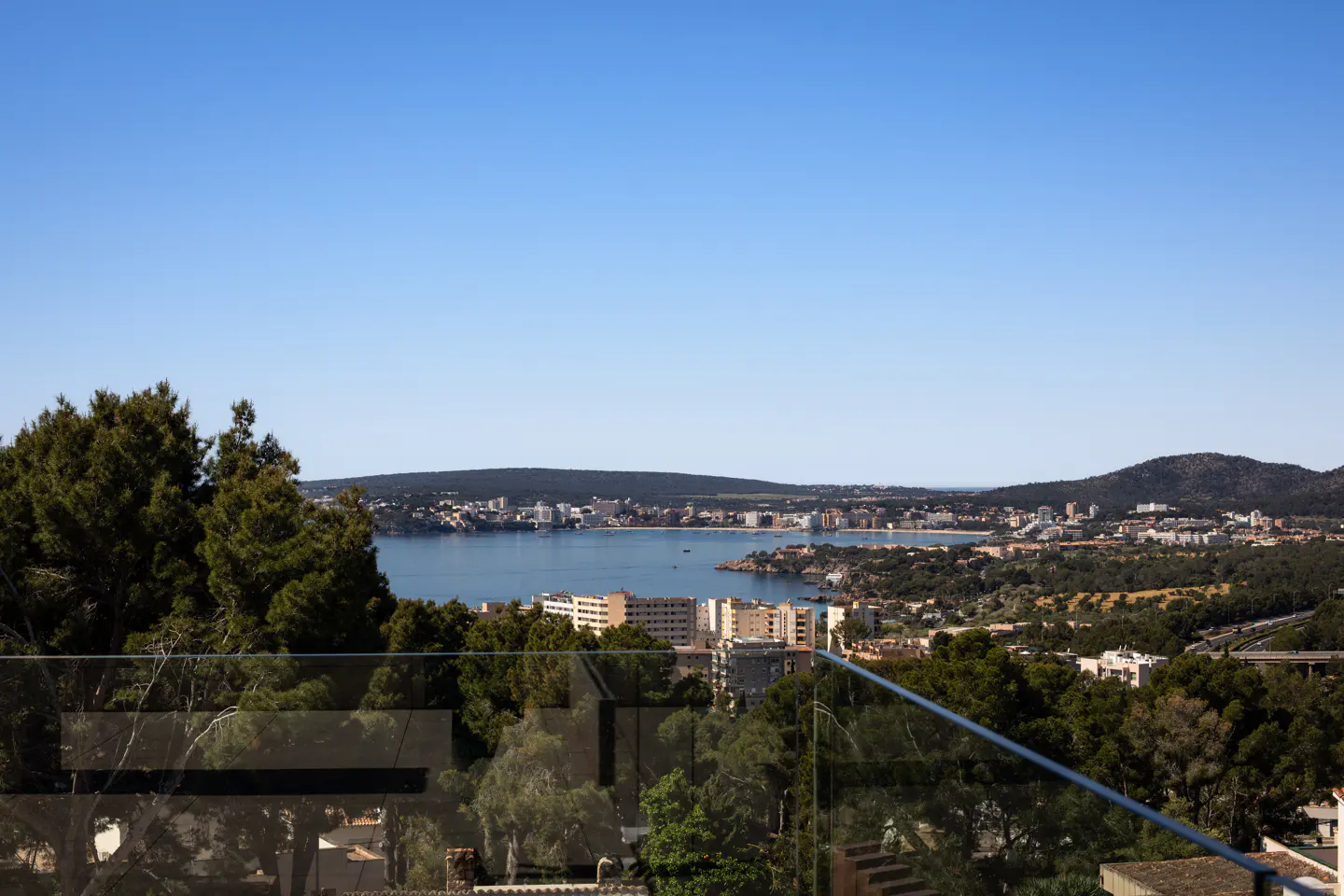View from a balcony with a glass railing overlooking a bay with buildings and trees under a blue sky.