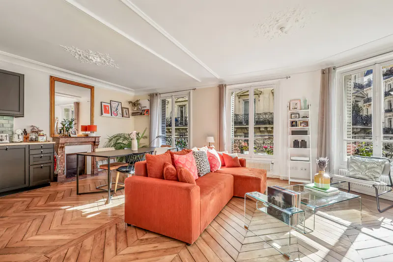 Bright living room with herringbone wood floors, orange sectional sofa, glass coffee table, and large windows overlooking city buildings.