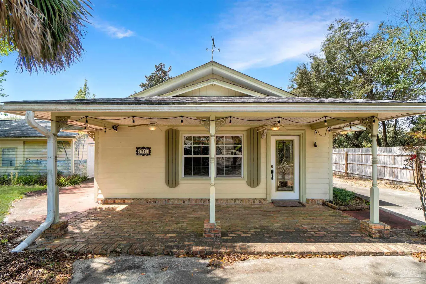 A pale yellow house with green shutters and a brick porch under a covered awning with string lights.