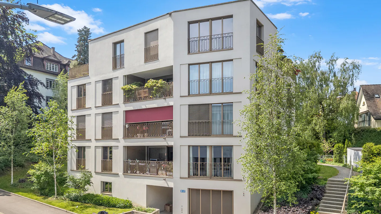 Exterior of a modern, white apartment building with balconies and green trees.