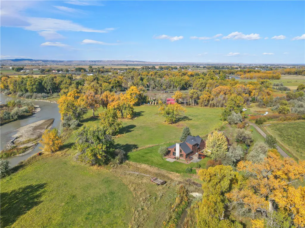 Aerial view of a brown house with a black roof, surrounded by green grass and trees with yellow leaves, under a blue sky.