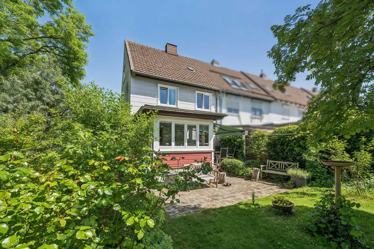 Backyard of a two-story house with a red-and-white facade, stone patio, wooden bench, and lush green garden.