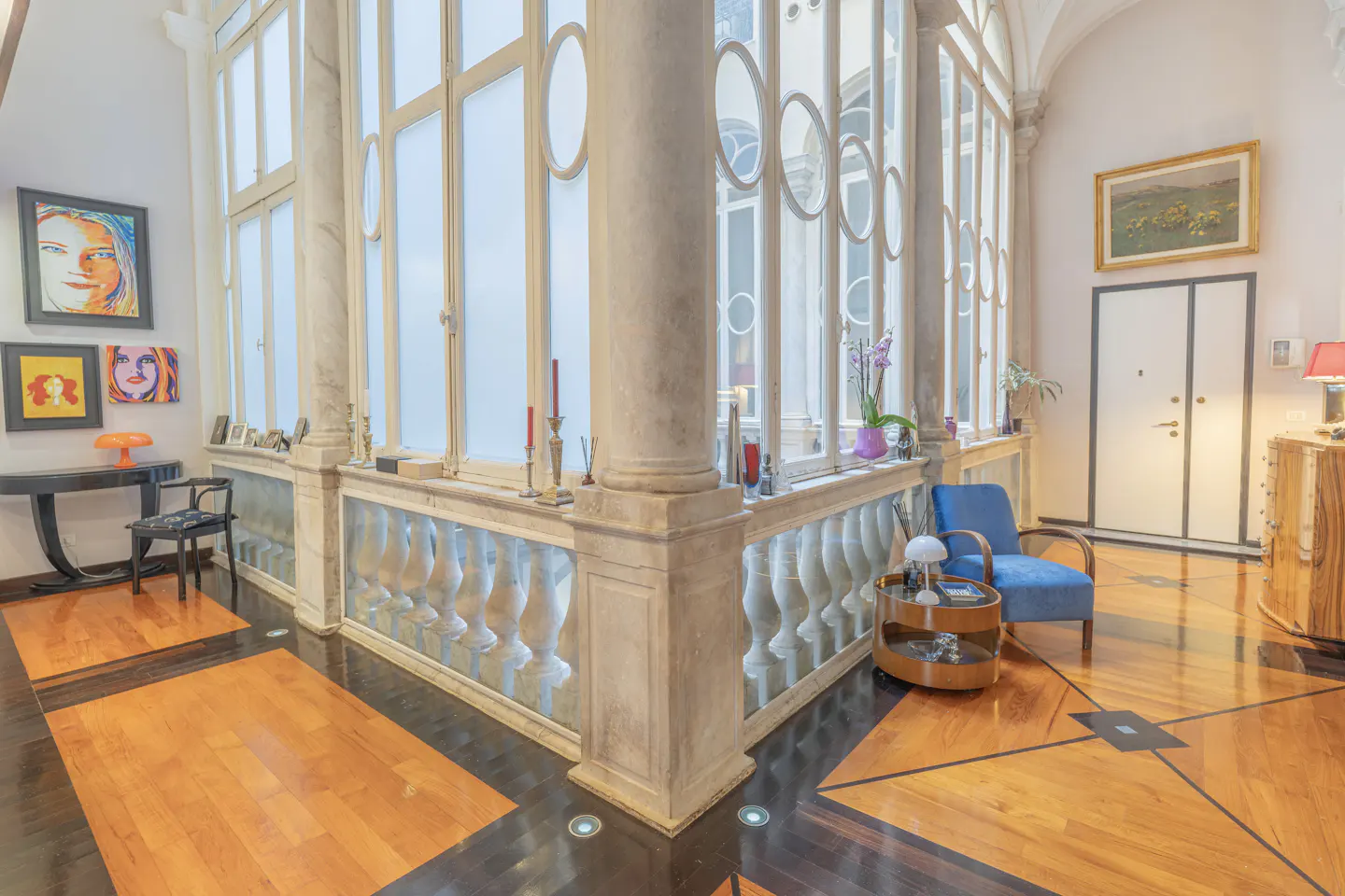 Bright hallway with marble columns, balustrade, and large windows. Art adorns the walls, and a blue chair sits near a round table.