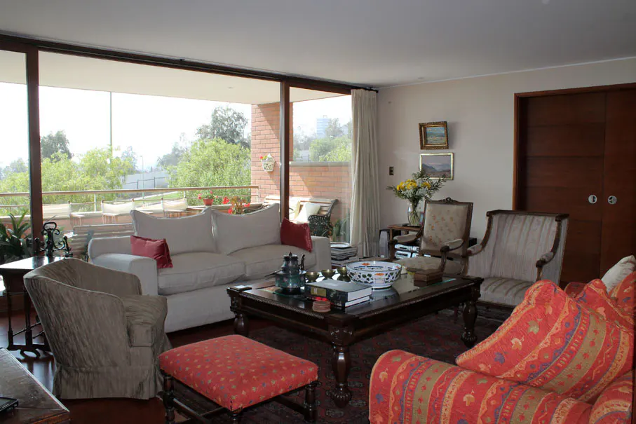 A living room with a white sofa, patterned chairs, and a dark wood coffee table. A balcony with greenery is visible through a large window.