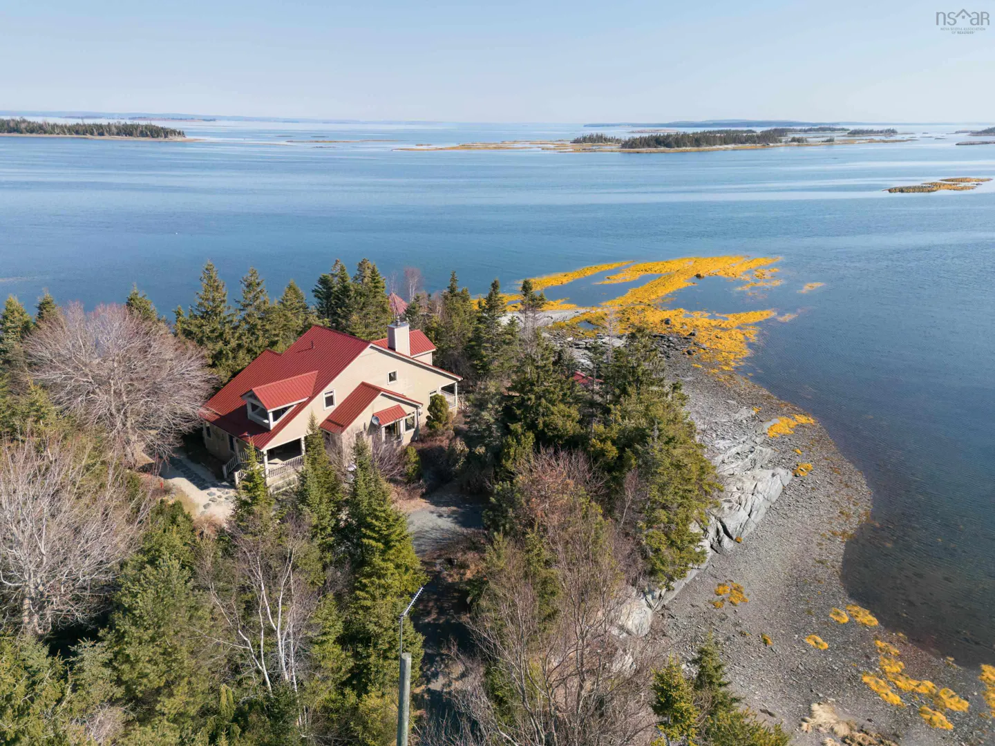 Aerial view of a tan house with a red roof, surrounded by trees, on a rocky coast next to a blue ocean.