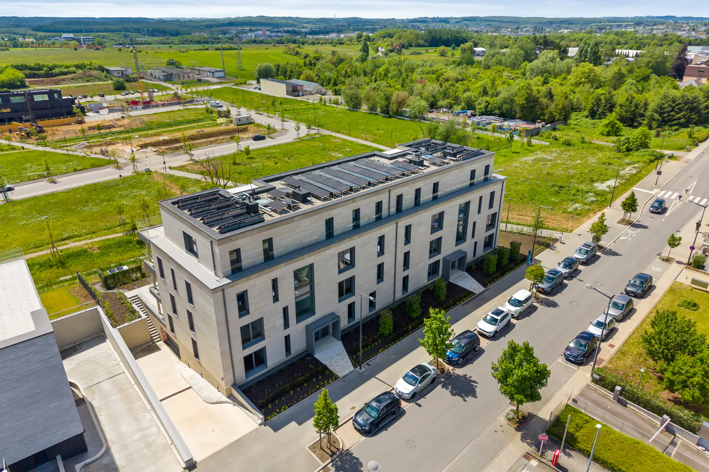 Aerial view of a modern, light gray building with solar panels on the roof, surrounded by green trees and parked cars.