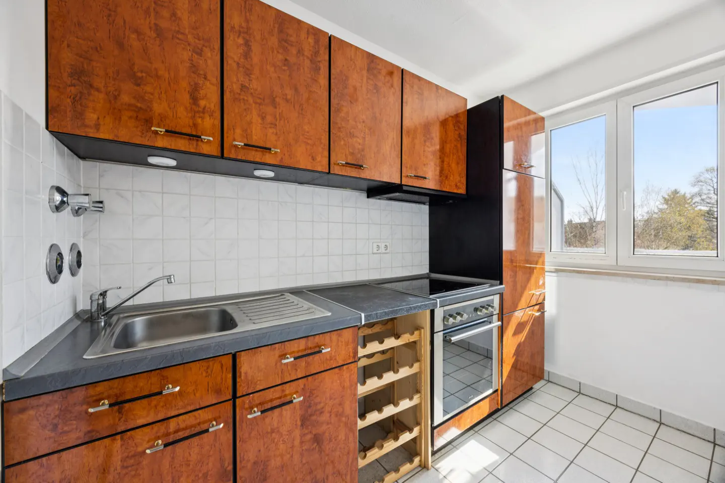 A kitchen with wood cabinets, a stainless steel sink, a black countertop, and a white tiled floor. A window is on the right.