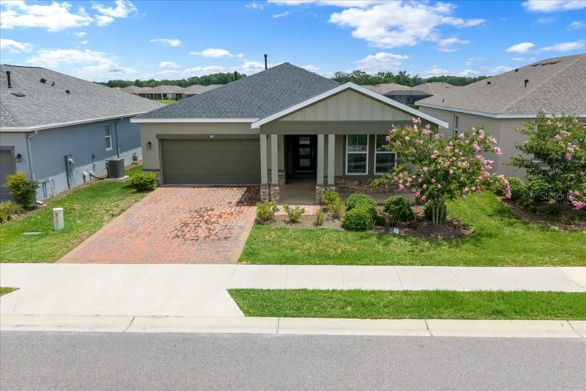A single-story house with a gray roof, olive garage door, brick driveway, and green lawn on a sunny day.