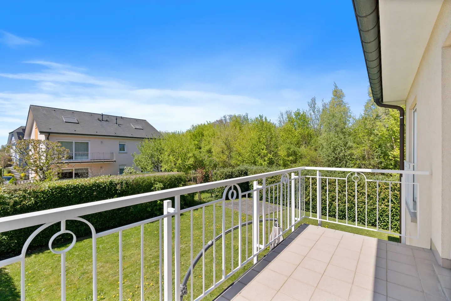 Balcony view with white railings overlooking a green lawn, trees, and a house under a blue sky.