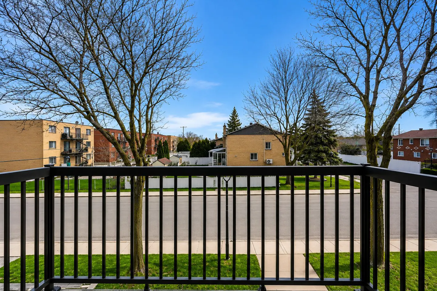 View from a balcony with black railings, overlooking a street with grass, trees, and brick apartment buildings under a blue sky.