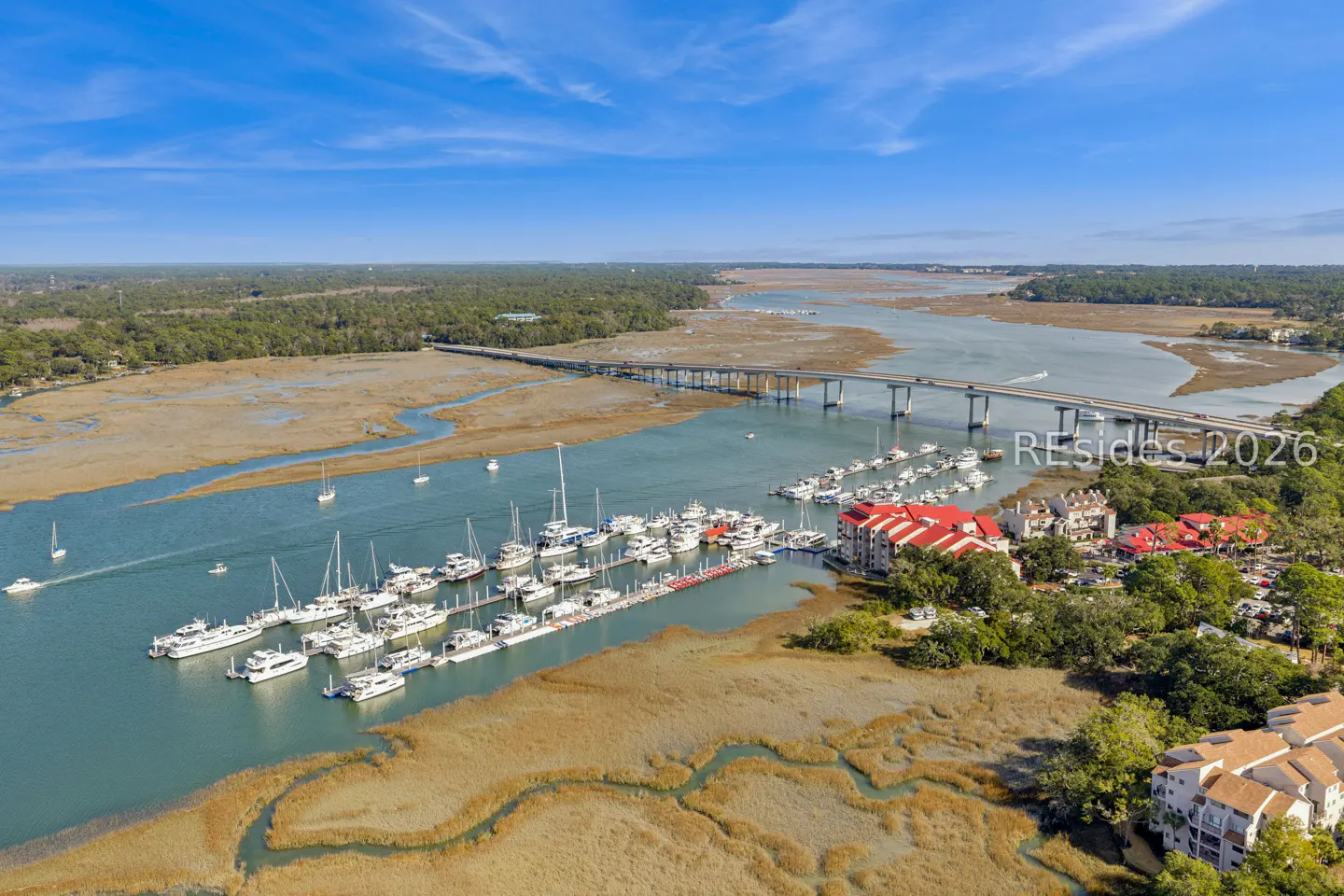 Aerial view of a marina filled with white boats, a bridge, and marshland under a blue sky.
