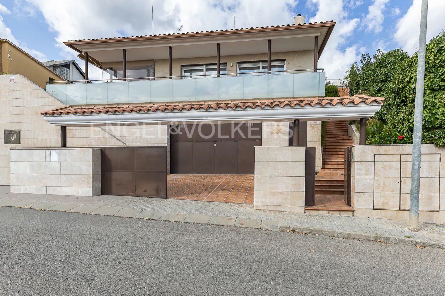 Two-story house with a balcony, brown garage door, and stone walls under a cloudy sky.