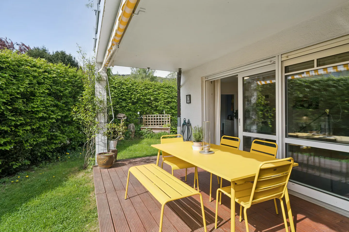 Outdoor patio with yellow table, chairs, and bench on a wooden deck. Green lawn and hedges surround the area. Sliding glass doors lead indoors.