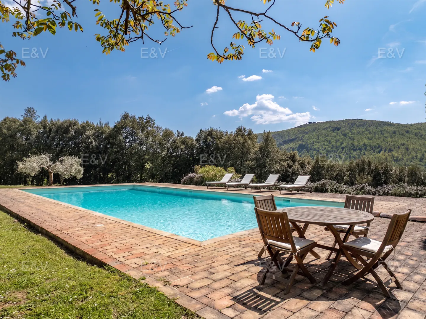 Outdoor pool with brick patio, table, chairs, and lounge chairs. Trees and a hill are in the background. Blue sky.