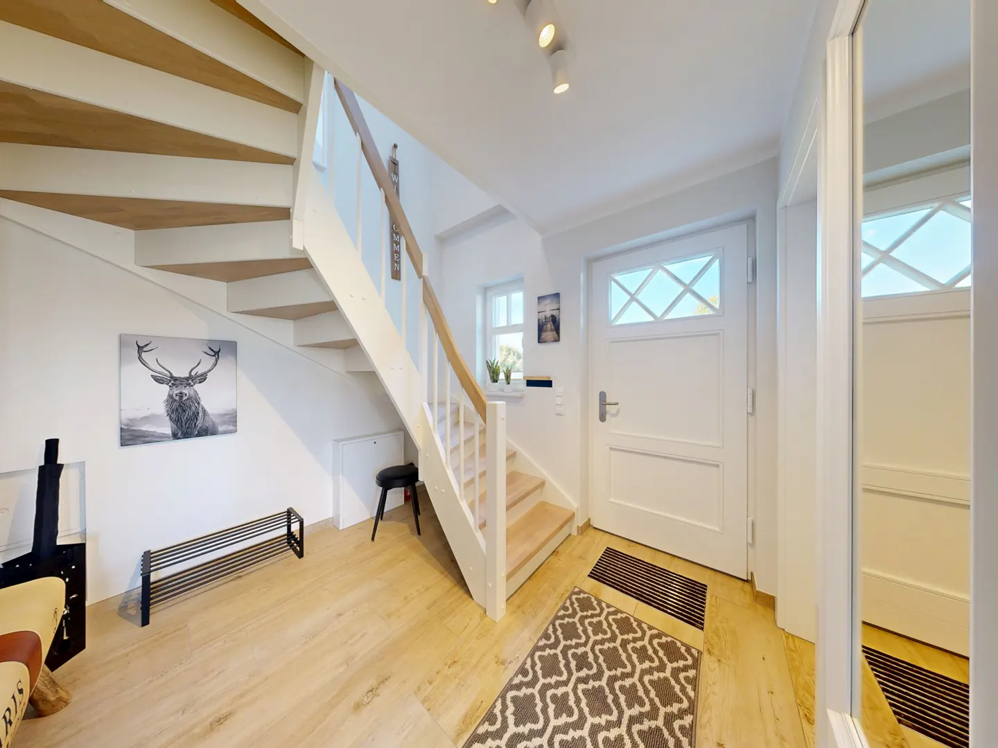 Bright foyer with white stairs, light wood floors, and a black and white deer print. A white front door and patterned rug complete the space.