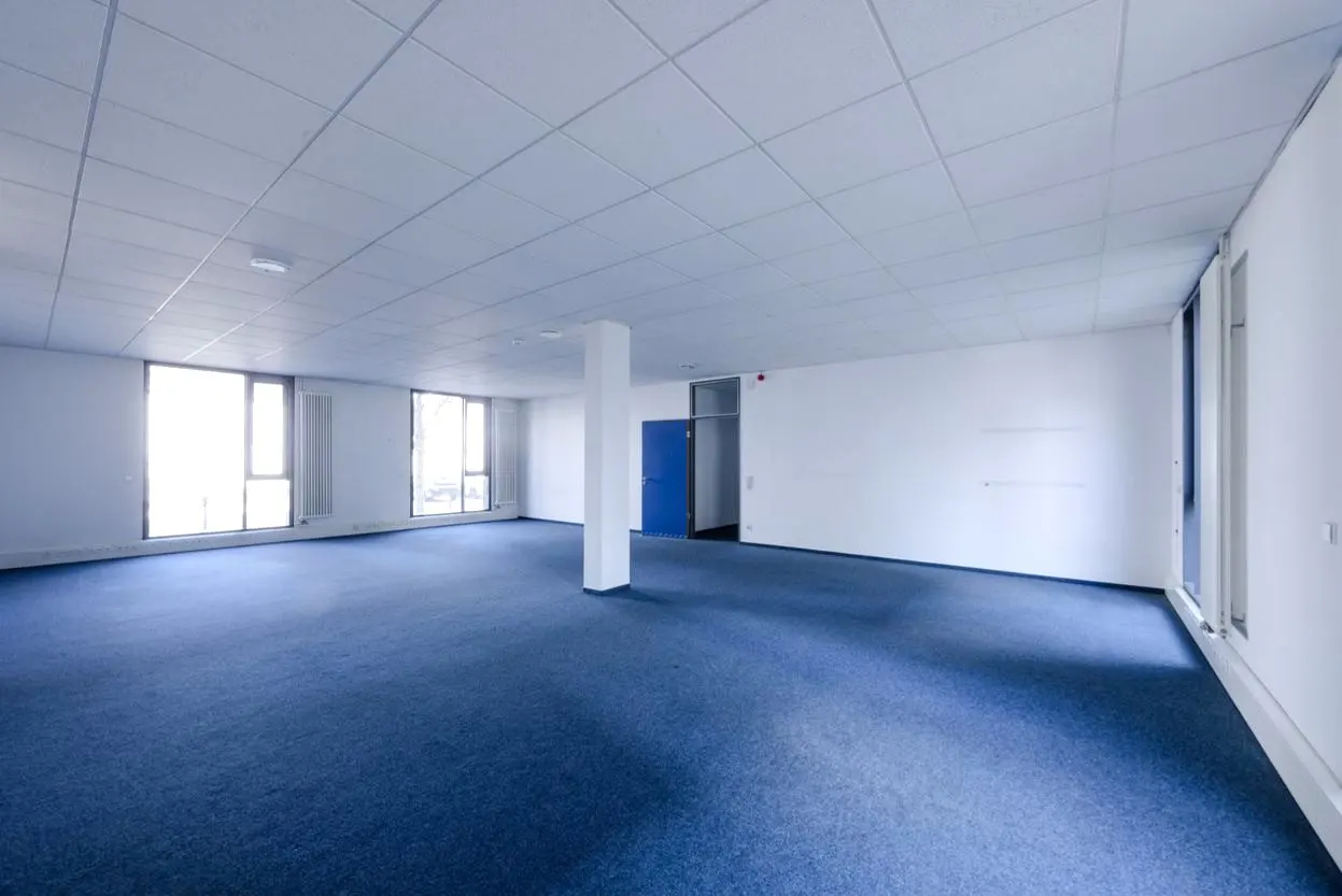 Empty office space with blue carpet, white walls, and a suspended ceiling. Windows line one wall, and a blue door is visible in the back.